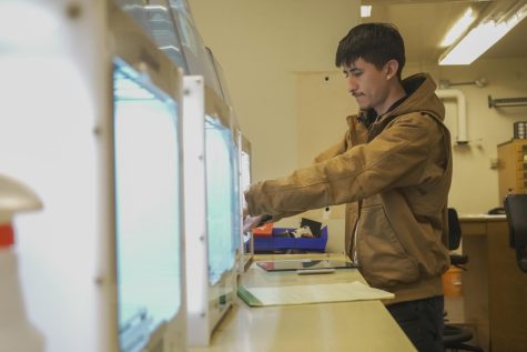 Gerardo Nunez, Junior, puts materials into the 3D printer in Hensill Hall at SF State on Tuesday, Feb. 14, 2023. Other 3D printing machines are found aside from the others aligned in a row. (David Jones / Xpress Magazine) 