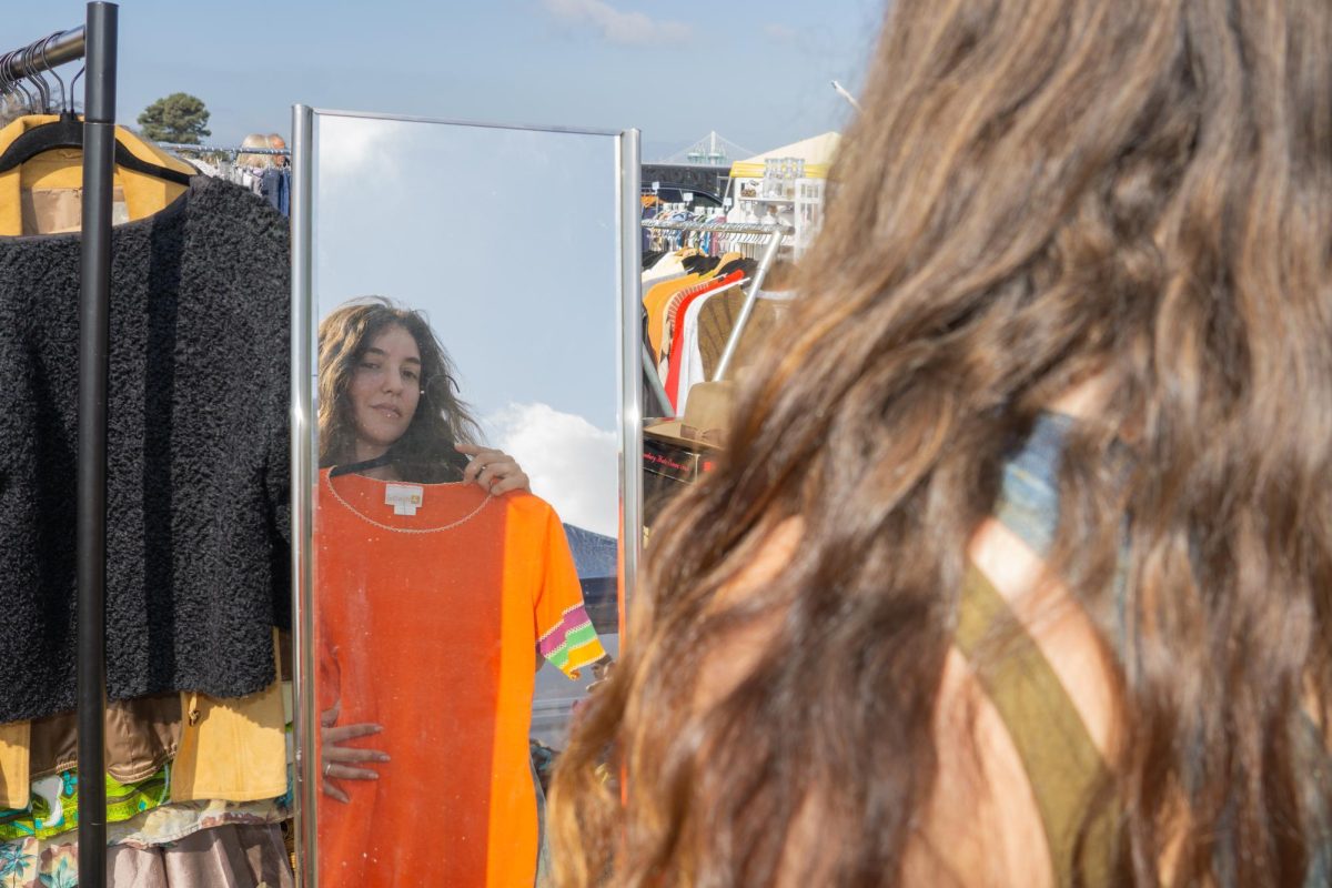 Current third year student at SFSU and vintage reseller who specializes in women’s clothing from the ’50s through ’70s, Maya Schraeder looks at a vintage orange dress through a mirror atthe Alameda Flea Market in Alameda, California. (Ryo Kojima/Xpress Magazine)