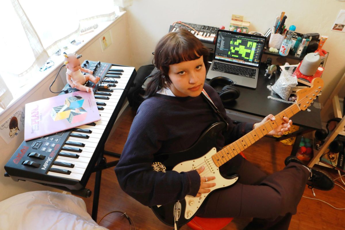 Chloe Little sits in their red rolling chair while they pose and hold their guitar in the corner of their bedroom studio, In San Francisco, Calif. on Thursday, Sept. 28, 2023. This is
the place where Little sits to record and produce their music. However, their space is also used for their poetry and art, the right corner of their desk is filled with piled up novels and composition books. (Leilani Xicotencatl/Xpress Magazine)